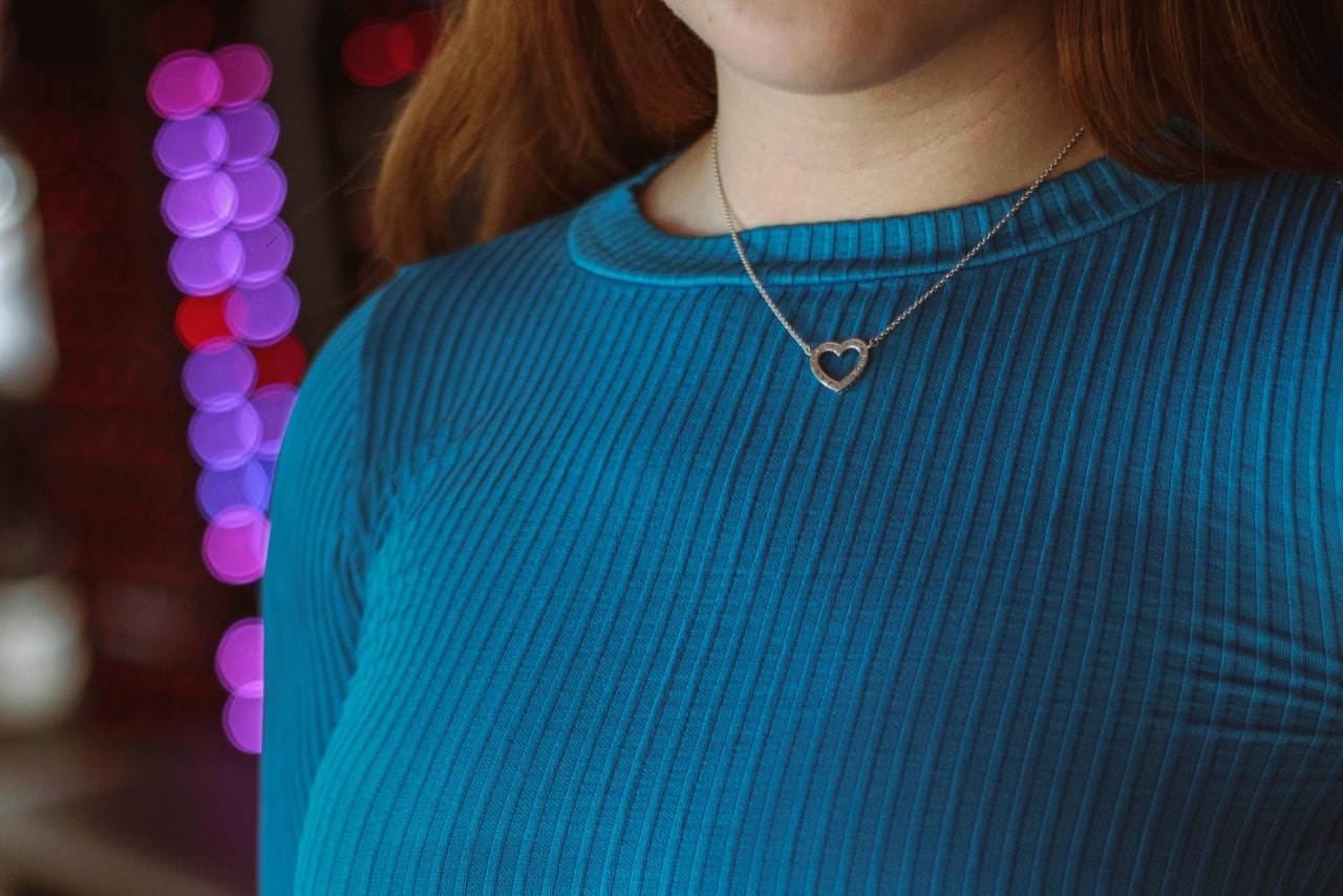 A close-up of a person wearing a blue ribbed top and a delicate platinum chain necklace with a small heart-shaped pendant.