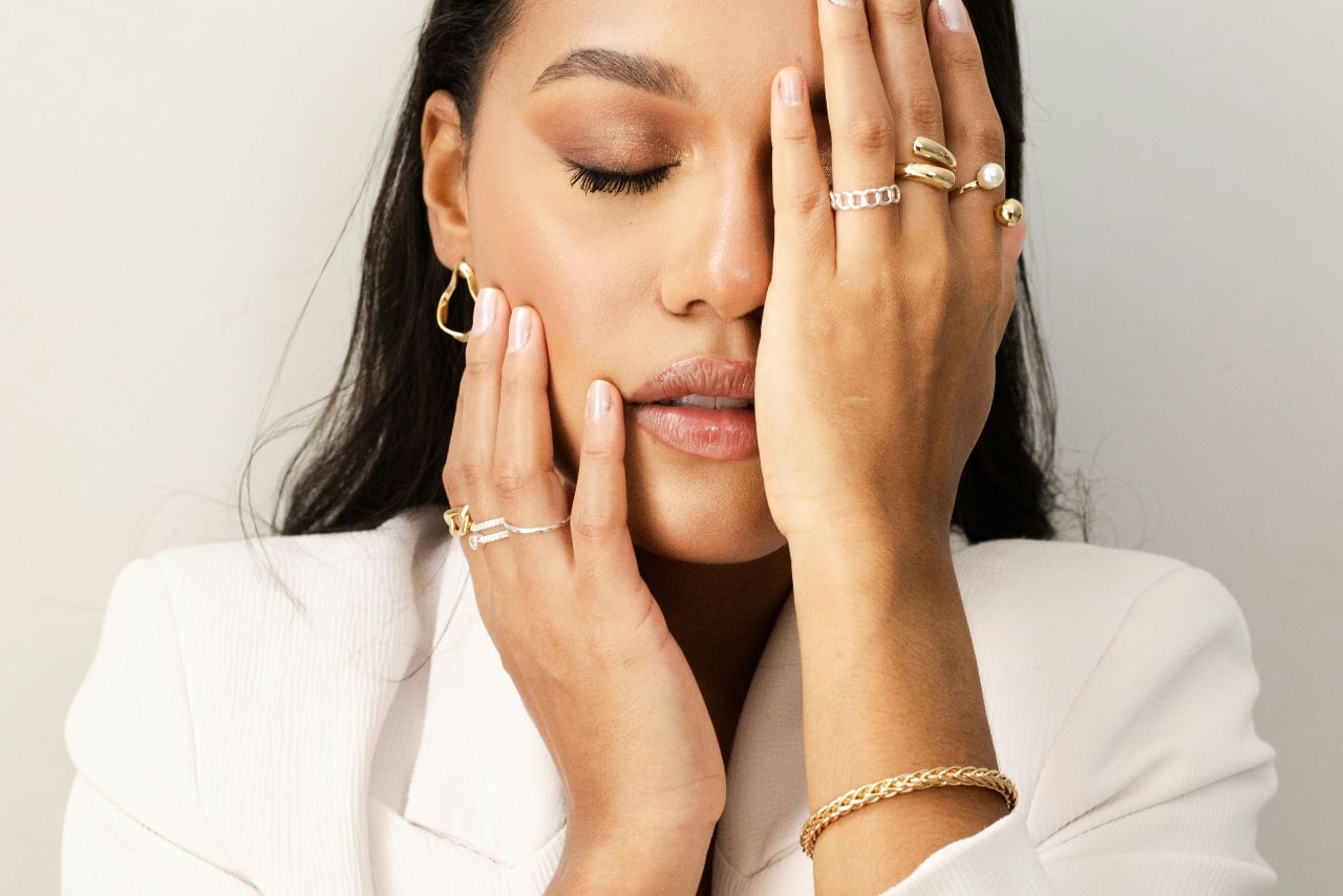 A woman covering half of her face with hand, showcasing various yellow and white gold rings.