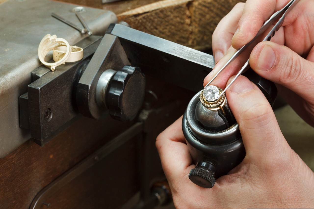 A close up of a jeweler placing with tweezers a round cut brilliant diamond in the halo setting, while the ring is being held by a special tool.
