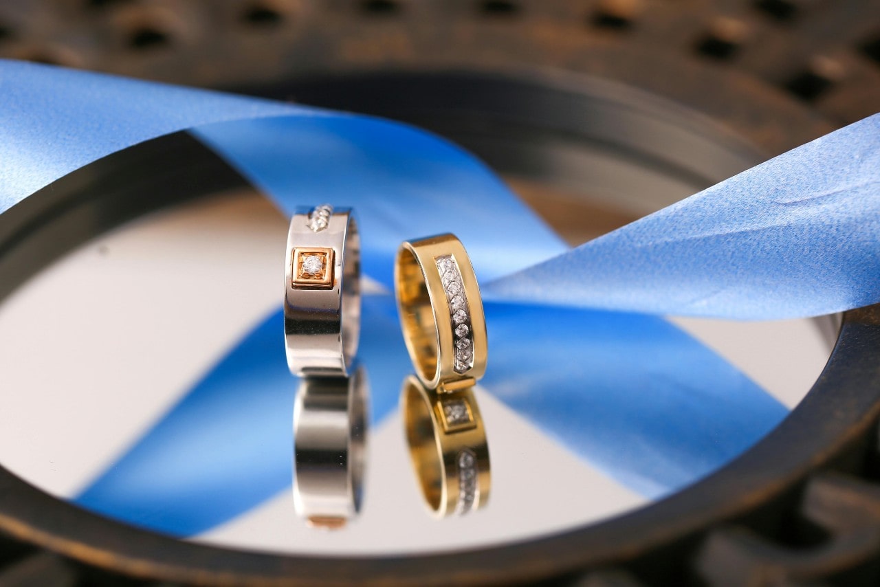 Two bold diamond wedding bands, one white gold and the other yellow gold, displayed atop of the reflective surface near the blue ribbon.