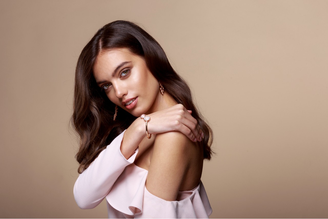 A woman with brown wavy hair in a pink shoulderless blouse showcases rose gold diamond jewelry. A woman with brown wavy hair in a pink shoulderless blouse showcases rose gold diamond jewelry.