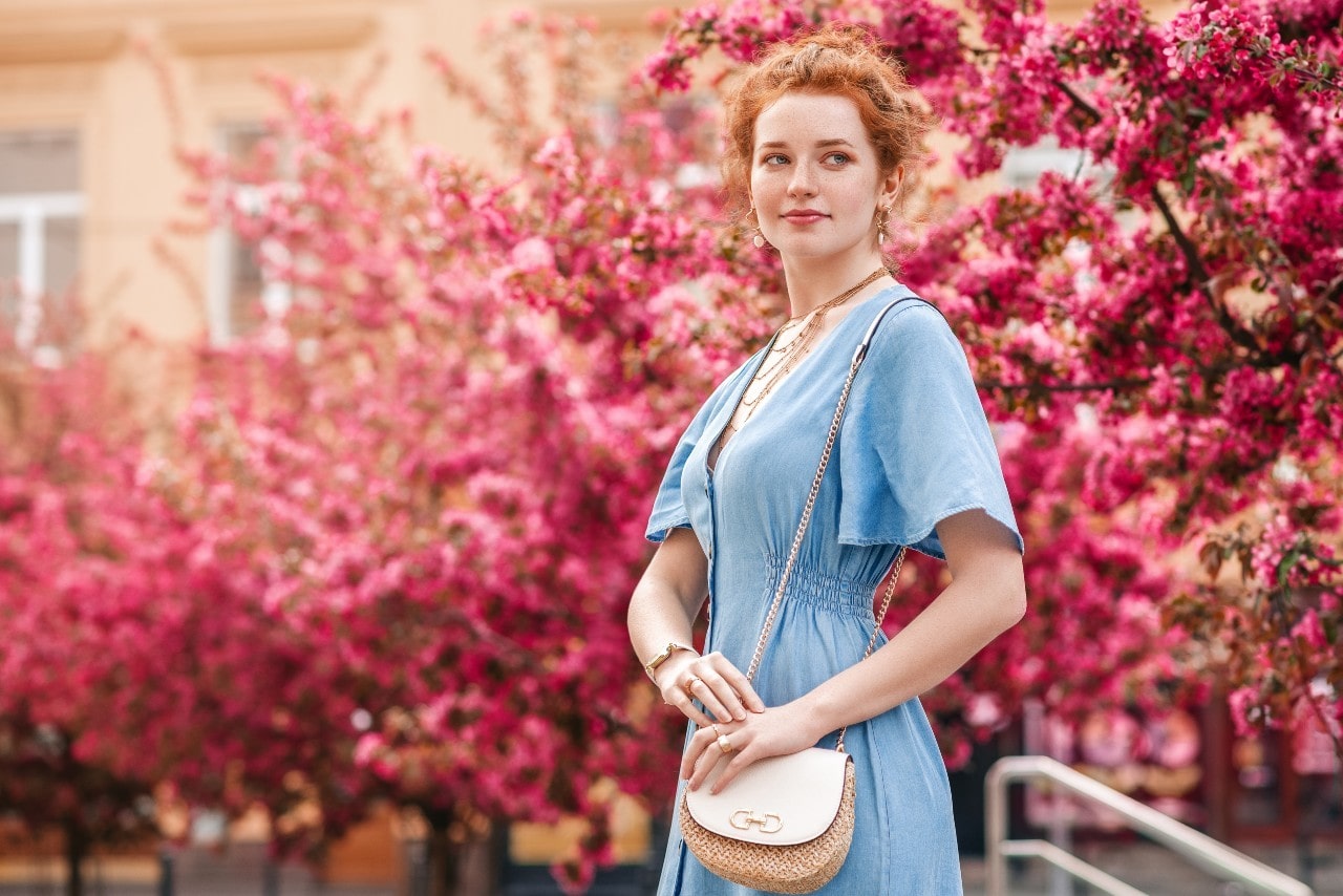 A woman in a blue dress and layered necklaces stands in front of blooming pink trees. A woman in a blue dress and layered necklaces stands in front of blooming pink trees.