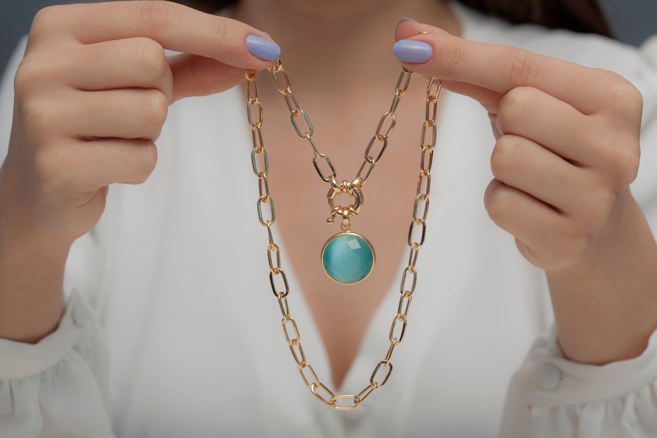 A close-up of a woman in a white blouse holding a yellow gold chain pendant with a round blue gemstone. A close-up of a woman in a white blouse holding a yellow gold chain pendant with a round blue gemstone.