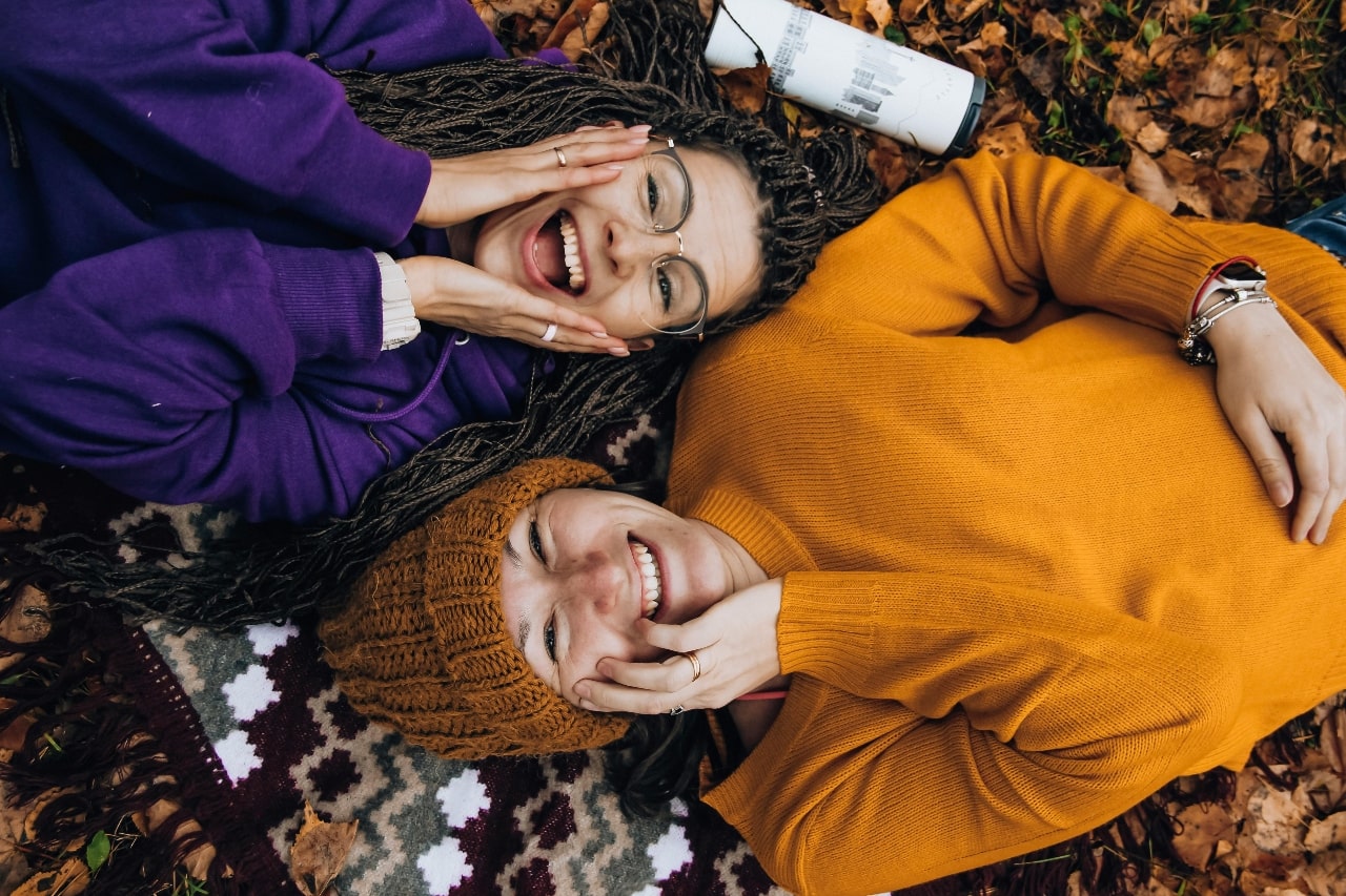 Two smiling friends lying on autumn leaves, showcasing casual stackable white gold rings and a charm bracelet. Two smiling friends lying on autumn leaves, showcasing casual stackable white gold rings and a charm bracelet.