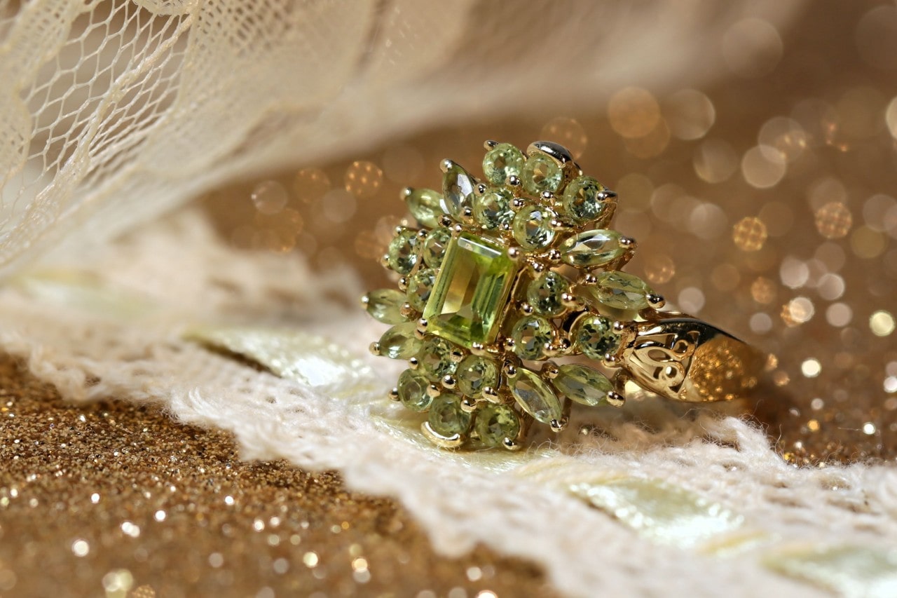 A yellow gold ring adorned with peridot gemstones rests on lace fabric with a sparkling, gold bokeh background.
