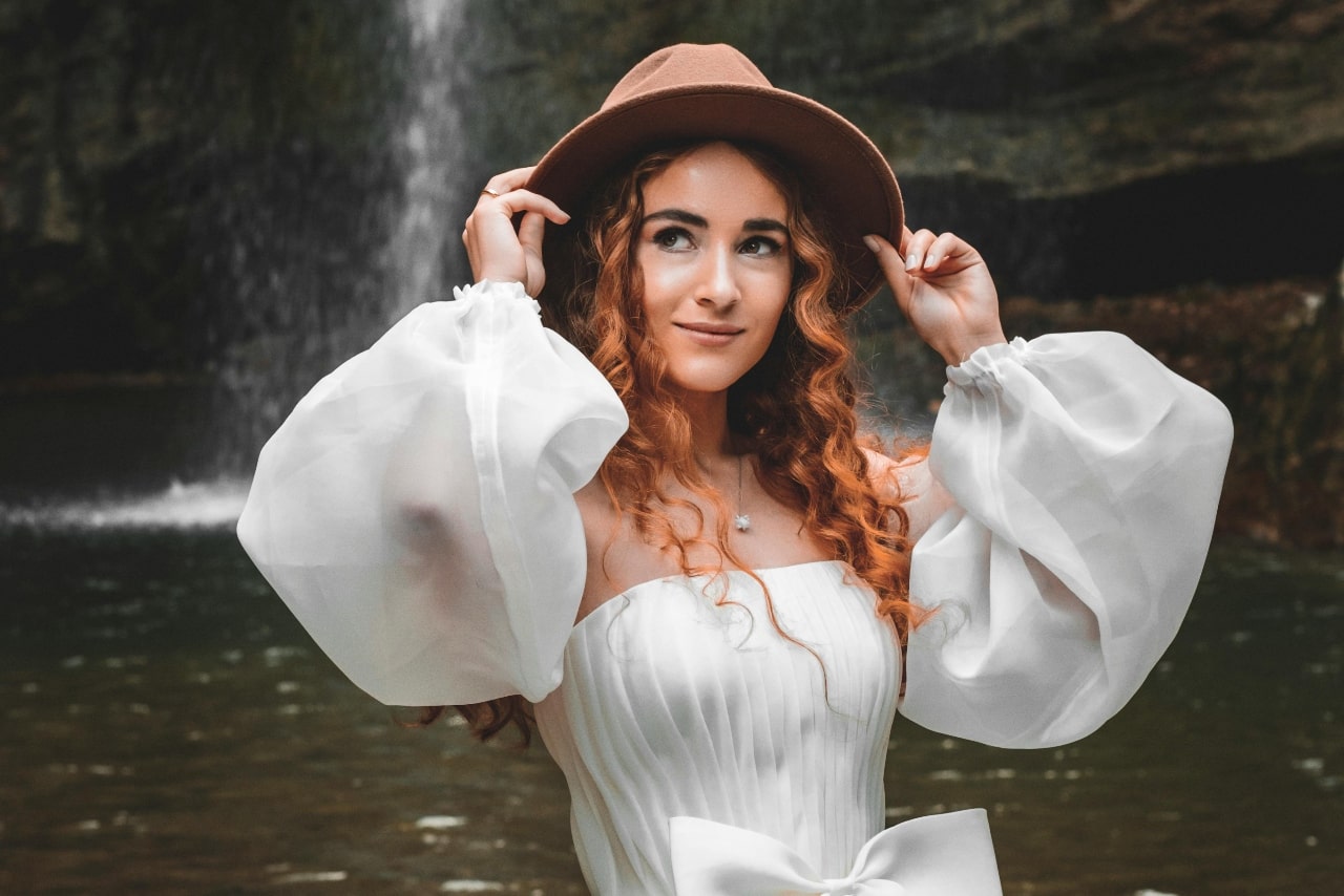 A young woman with curly hair in a white dress, wearing a diamond pendant and wide-brimmed hat stands by a waterfall.