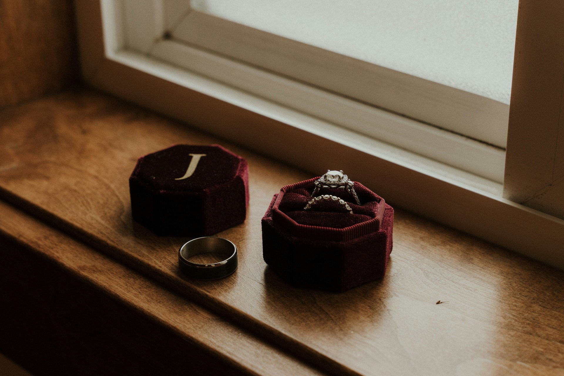 An open red velvet box with the set of diamond engagement ring and wedding band inside, while tungsten men’s wedding band lay nearby on a wooden surface.