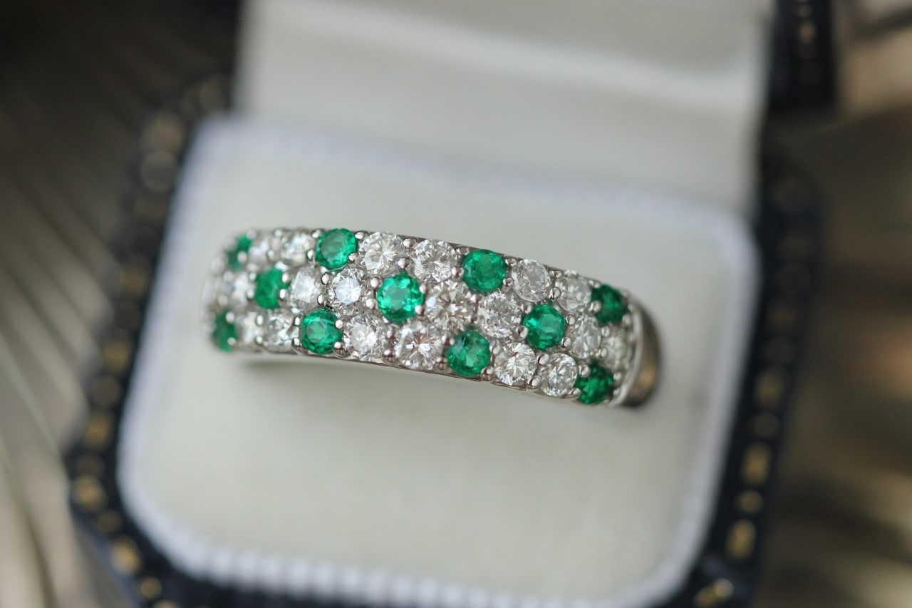 A close up of a three row wedding band with alternating round cut diamonds and emeralds inside a blurred jewelry box.