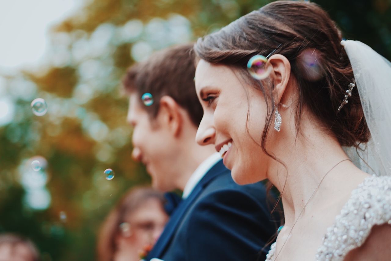A side view of a bride and her groom in the background, with bubbles floating nearby. A side view of a bride and her groom in the background, with bubbles floating nearby.