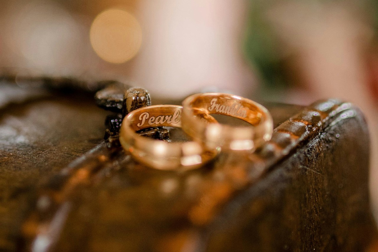 A pair of yellow gold wedding bands atop a leather chest, with the names “Pearl” and “Franklin” engraved inside. A pair of yellow gold wedding bands atop a leather chest, with the names “Pearl” and “Franklin” engraved inside.