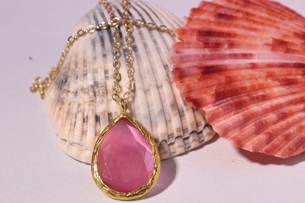 a yellow gold pendant necklace featuring a pink gemstone lying over two shells