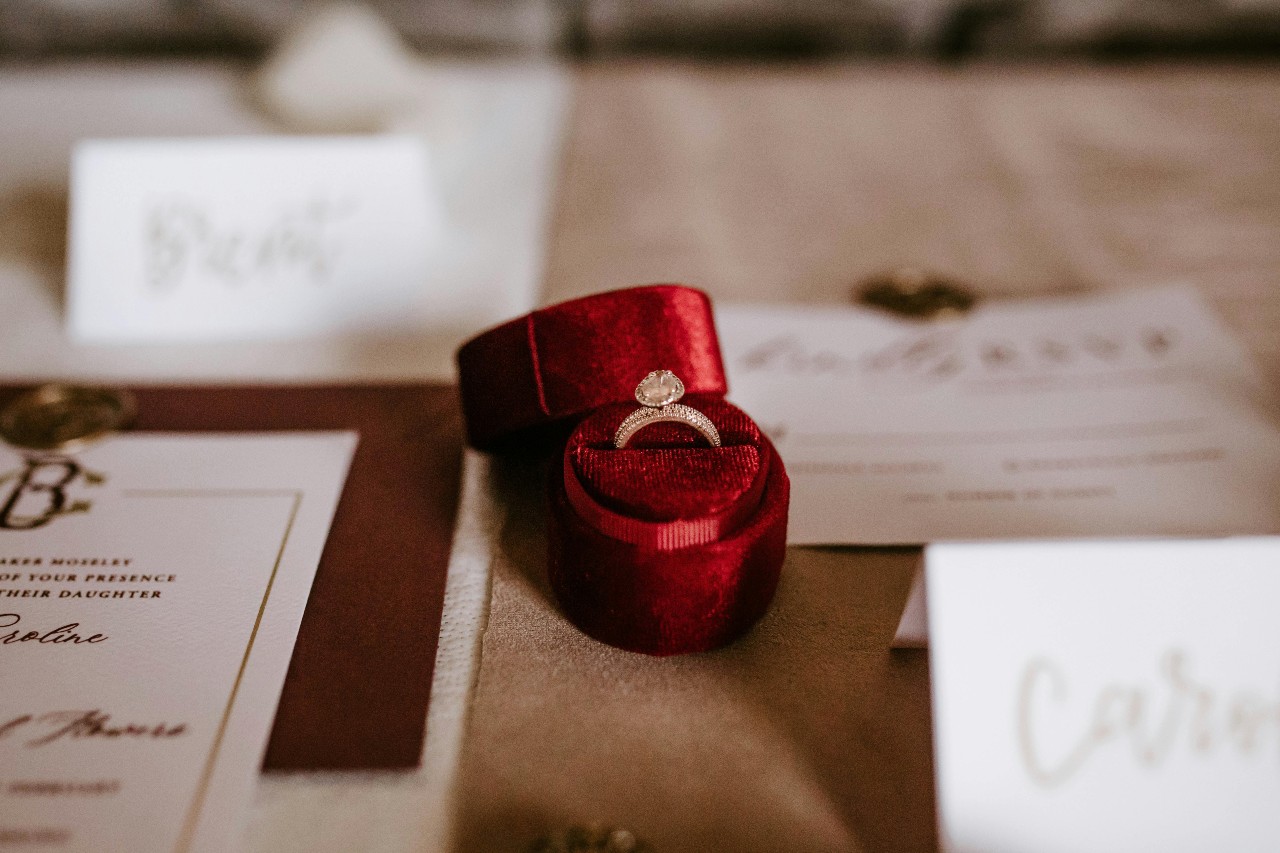 An oval-cut side stone ring sits in a red velvet ring box among wedding invitations.