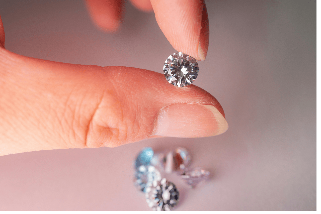 A close-up of a person’s hand holding a round cut diamond between their fingers with more diamonds on the surface below.