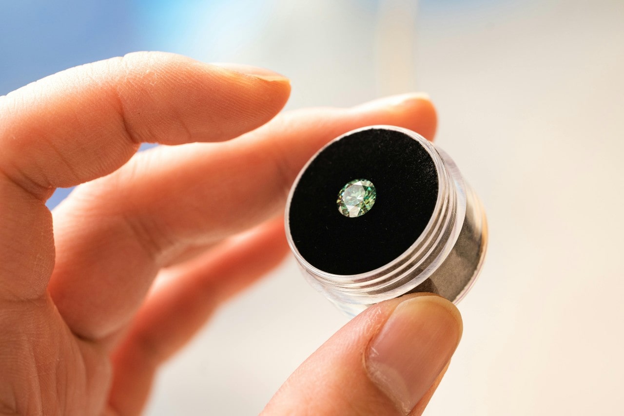 A close-up of a person holding a metal case with a green round cut diamond inside.
