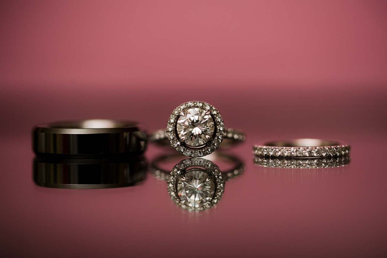A men’s wedding band, halo engagement ring, and diamond wedding band on a reflective surface with a magenta background.