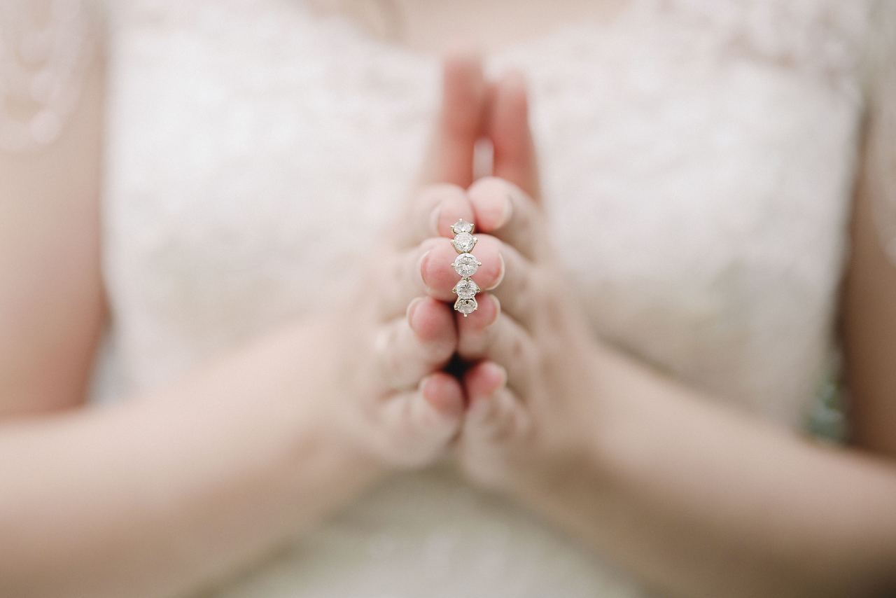 A woman putting her hands together with a diamond wedding band in between them, her wedding gown blurred in the background.