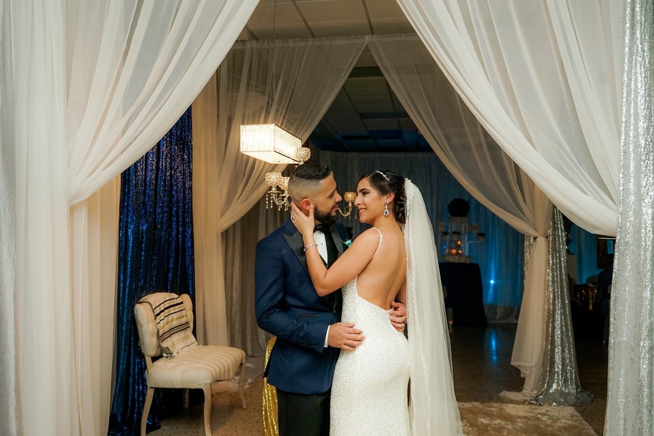 A bride and groom about to kiss as they stand in their wedding reception venue decorated with lots of sparkly curtains. A bride and groom about to kiss as they stand in their wedding reception venue decorated with lots of sparkly curtains.