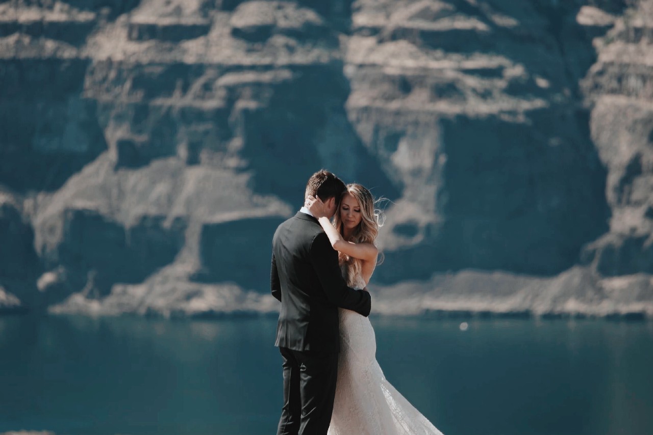 A couple dressed in their wedding attire embracing on a high cliff, a large rock formation and water behind them. A couple dressed in their wedding attire embracing on a high cliff, a large rock formation and water behind them.