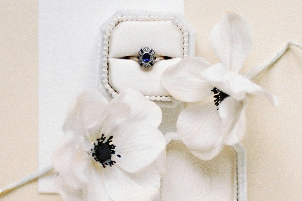 A delicate vintage-style engagement ring with a deep blue sapphire center stone and intricate diamond detailing, displayed in a white velvet ring box with white flowers in the background