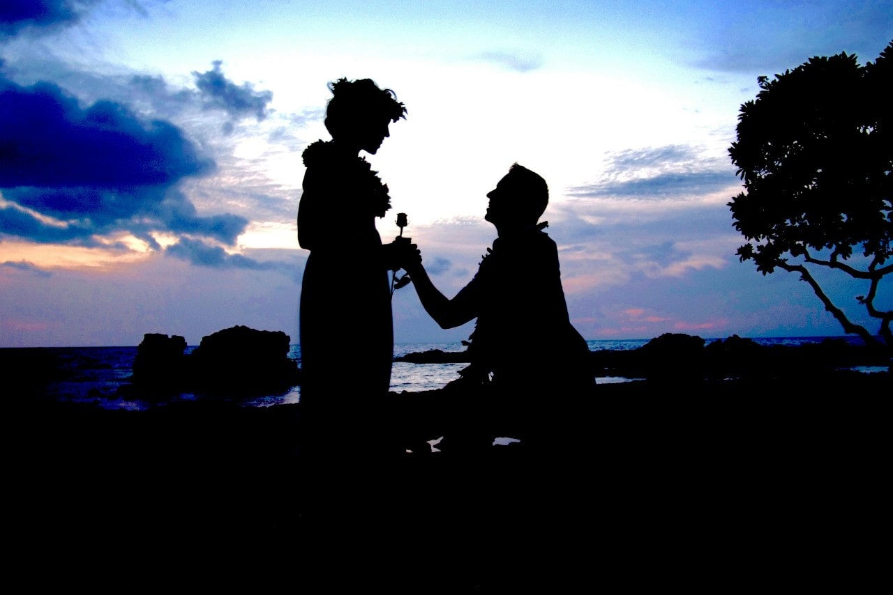 A romantic silhouette of a man proposing to a woman on a beach at sunset, with the ocean and a dramatic sky in the background.